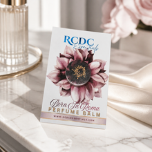 RCDC Essentials perfume balm in a round glass jar with a floral label, displayed on a marble surface with a silk ribbon and a perfume bottle in the background.